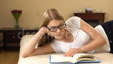 美丽的年轻女子穿着白色的t恤和眼镜，轻松地躺在沙发上，享受着看书的乐趣。
