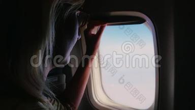 一个年轻的女人关上一扇飞机窗户，<strong>看</strong>着<strong>窗外</strong>。 黑暗的框架被照亮的开口