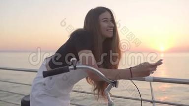 年轻漂亮的女人在日出或日落时使用智能手机，在海边骑老式自行车