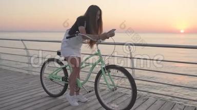 年轻漂亮的女人在<strong>日</strong>出或<strong>日</strong>落时使用智能<strong>手机</strong>，在海边骑老式自行车