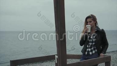 可爱的金发女人穿着黑色皮夹克在海边喝咖啡