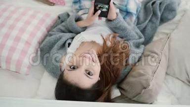 年轻漂亮的女人躺在床上，在智能手机上看照片。 女孩看着相机微笑。