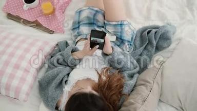 年轻漂亮的女人躺在床上拿着手机。 女孩在智能手机上看照片，喝果汁。