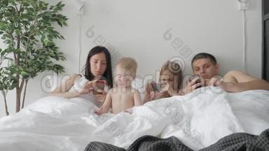 年轻的家庭使用小工具躺在白色的床上。 概念现代家庭周末在家。 当代科技。