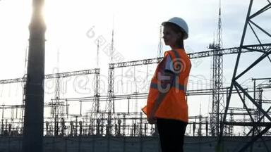 一位戴着白色头盔、穿着橙色防护背心的女工程师在检查一个有电线和电力的发电站