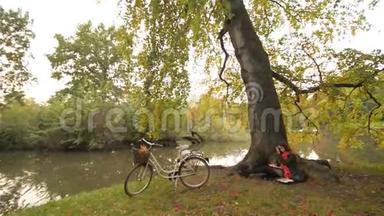 美丽的学生女孩放松在秋天公园看书，独自坐在树下，自行车在背景，阳光
