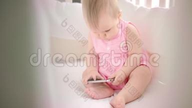 婴儿床上有电话的小女孩。 幼儿在手机上观看动画片