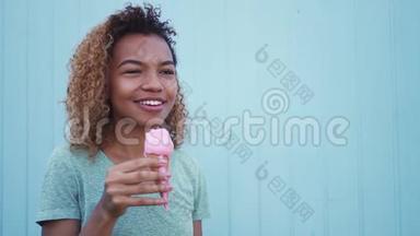 年轻的黑人女孩在蓝墙背景下吃粉色融化冰淇淋