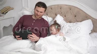 英俊的父亲和他的小女儿躺在床上，使用电子设备。 小辫子的女孩子