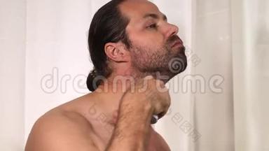 英俊的年轻人留着长发，在浴室里用<strong>修剪器</strong>刮胡子。 特写镜头。