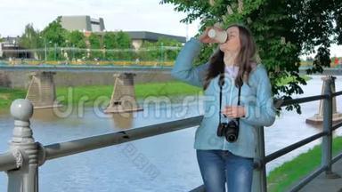 美丽的年轻女孩摄影师，带着专业的相机，在海滨、河边和河边的栏杆旁喝咖啡。
