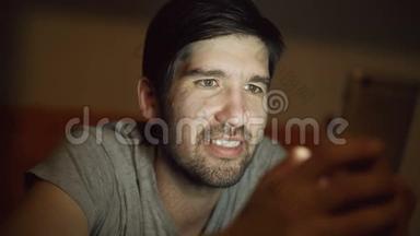 年轻的微笑男子使用智能手机浏览社交媒体，晚上躺在家里的床上