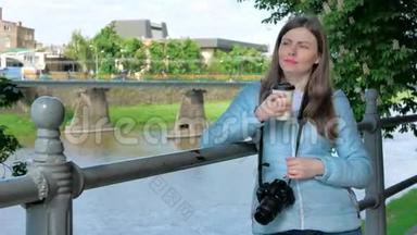 美丽的年轻女孩摄影师，带着专业的相机，在海滨、河边和河边的栏杆旁喝咖啡。