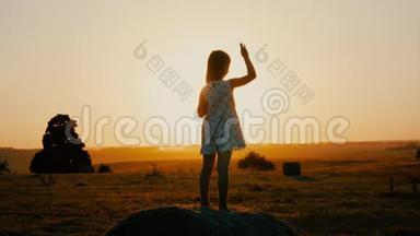 小女孩在日落的草地上挥手。 日落时分在田野里快乐的漂亮女孩。