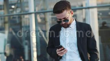 一个戴太阳镜的年轻胡子男子站在机场候机楼旁，用他的<strong>手机旋转</strong>。 商务
