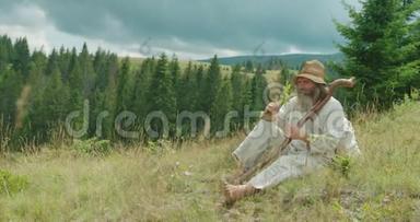 穿着旧衣服的老人坐在山上的草地上闻着花。 横向视野