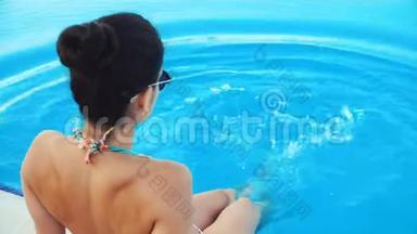 年轻的黑发女人，穿着比基尼，戴着太阳镜，坐在游泳池里，在阳光明媚的夏日泼水