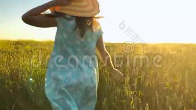美丽的女孩戴着黄色的帽子在日落的天空上的绿色麦田上奔跑。 自由概念。 日落时的麦田