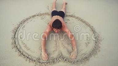 年轻开朗的人仰面躺在沙滩上，在海滨潮湿的沙滩上画着一个心的象征。 慢动作