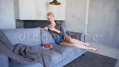 迷人的金发女郎使用智能手机，尝试成熟的草莓，坐在客厅的软沙发上，灰色的墙壁在客厅里