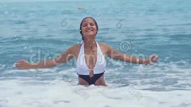 穿着漂亮比基尼的快乐年轻女子坐在海边的水里，举起手来，泼水，在阳光明媚的日子里微笑