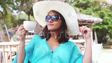 年轻优雅的黑发女人，带着帽子和太阳镜度假，坐在海滩咖啡馆，欣赏海景。