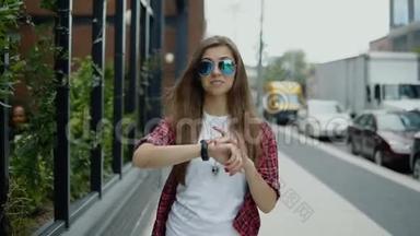年轻貌美的女士戴着太阳镜，穿着时髦的衣服，在街咖附近散步，查看时间