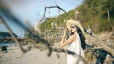 戴着太阳镜和帽子的年轻女子微笑着在美丽的热带海滩的树旁摆姿势