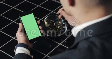 年轻的商人使用带有绿色屏幕触摸板的smarthpone，并在咖啡馆打手势接近。 一个拿着手机的家伙，敲