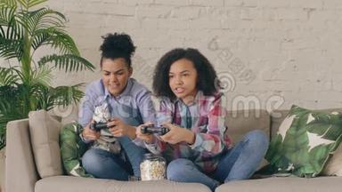 慢动作两个混合种族卷曲女孩朋友坐在沙发上玩控制台电脑游戏与游戏本和乐趣