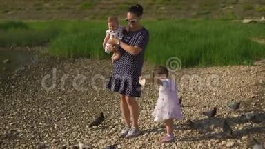 年轻的母亲带着她的小女儿在河边喂天鹅和小鸭子，小鸟在河边吃面包，穿着点缀的裙子
