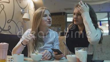 两个漂亮的年轻女孩坐在咖啡馆里，吃着看着手机屏幕。