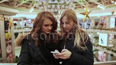 两个少女在<strong>商城</strong>，饶有兴致的看着手机屏幕.. 他们穿着夹克。