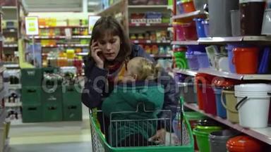 一个年轻的女人带着一个孩子在超市选择<strong>商品</strong>。 妈妈在手推车上驾驶一个小女孩。 考虑一下货物