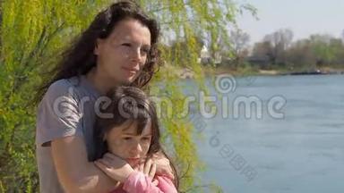 妈妈在河边拥抱她的女儿。 一个小女孩和她妈妈在水边。 家庭在露天。