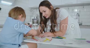 年轻漂亮的妈妈和儿子坐在厨房的桌子旁用彩色铅笔画画