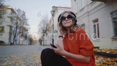 可爱的少女听耳机音乐，都市风格，时髦的时髦少女坐在城市街道的人行道上