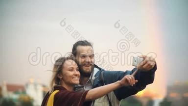 年轻的幸福<strong>夫妇</strong>在背景上拍摄彩虹的<strong>自拍照</strong>片。 微笑的男人和女人使用智能手机。