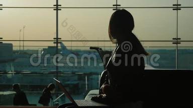 在机场<strong>候机</strong>楼用笔记本电脑工作的年轻女子。 在等我的航班。 在一个大的背景下剪影