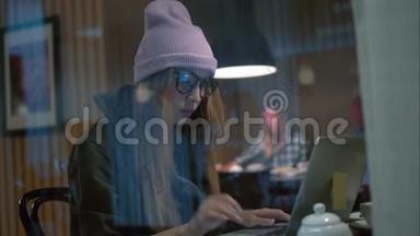 一个戴着眼镜和帽子的时髦女人坐在咖啡馆里，用笔记本<strong>电脑打字</strong>。 从窗口观看