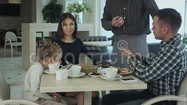 快乐的母亲、父亲和小女孩吃晚饭，请服务员用智能手机拍照。