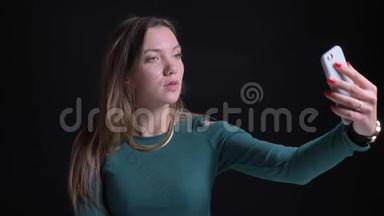 年轻的黑发白种人女孩在黑色背景下使用智能手机制作自拍照片的肖像。