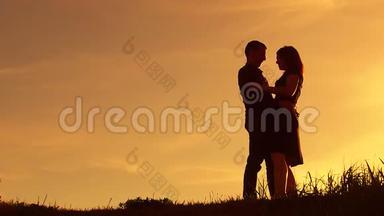 剪影男在日落时围着女孩玩拥抱。 男人和女人的剪影在夕阳下缓慢的运动。 恋爱中的情侣