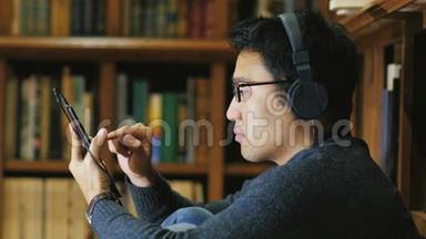 一个戴眼镜的亚洲年轻人喜欢这款平板电脑。 坐在图书馆书架的背景上..