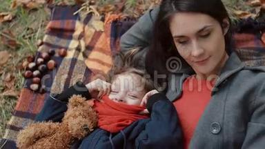 美丽的母亲和她可爱的小女儿躺在格子布上，在秋天公园玩枫叶