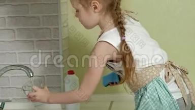 漂亮的小女孩穿着围裙在明亮的厨房里洗了一个杯子，帮助父母