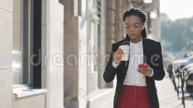 一位年轻的非洲裔美国女商人穿着西装，在城市里走来走去，喝着咖啡，用着