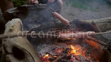徒步旅行者的朋友们<strong>围坐</strong>在篝火旁，用棍子炸香肠。 晚上在森林里户外露营野餐.. 高清高清