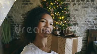 年轻的非洲裔美国女孩在<strong>家中</strong>圣诞树附近使用智能手机摄像头在线聊天