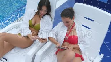 两个拿着平板电脑和电话的年轻女子坐在游泳池里